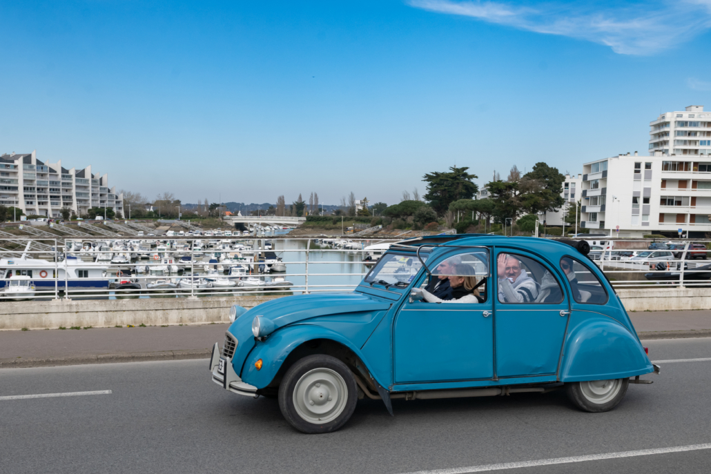 Participants à bord d'une Citroën 2CV bleue lors d'un rallye touristique en équipe près d'un port de plaisance.