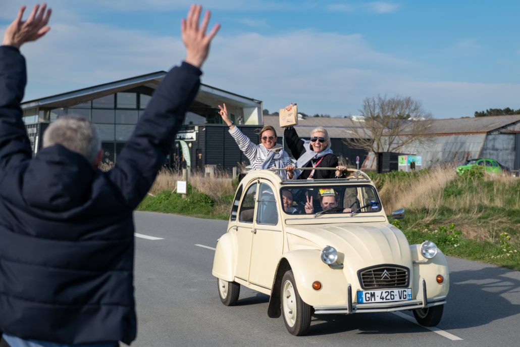 Équipage saluant les autres participants depuis une Citroën 2CV décapotable lors d'un challenge de cohésion.