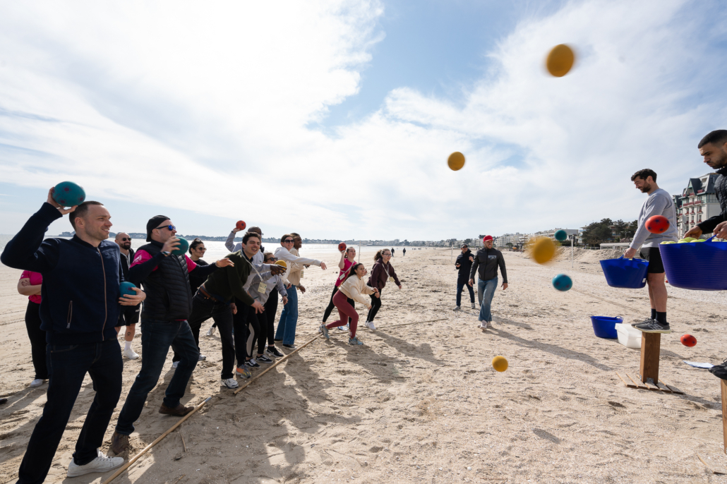 Groupe de collaborateurs participant à un défi collectif de lancer de balles sur la plage.