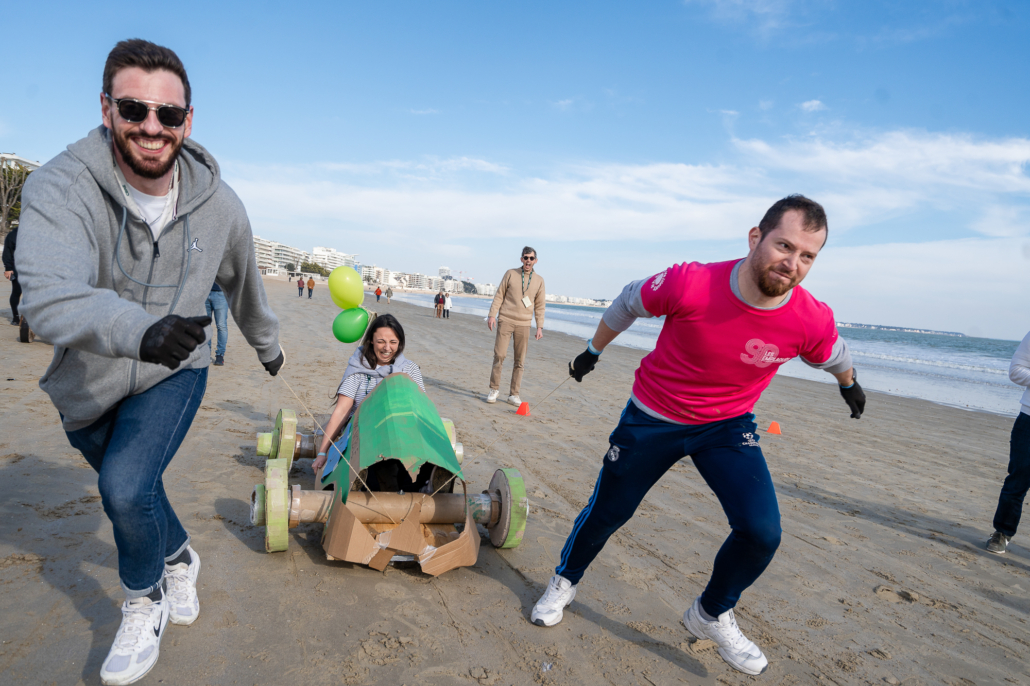 Deux participants tirent une caisse à savon sur le sable tandis qu'une collègue prend place à bord lors d'une animation de team building.